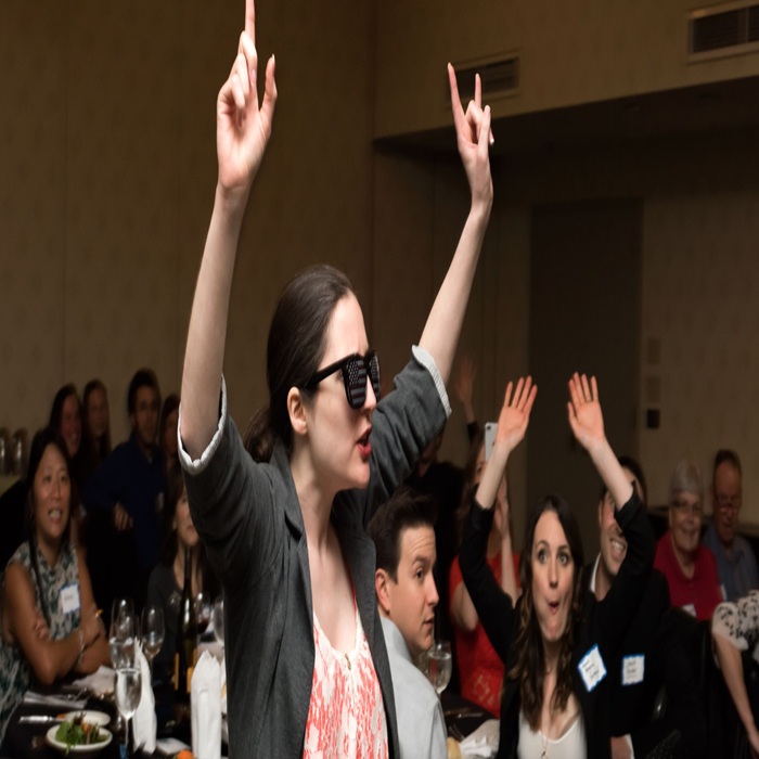 Woman in a blazer and pink top raises both hands at a seated event, wearing novelty 3D glasses, with a cheering crowd in the background.