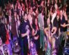 Crowded indoor party scene with people dancing and socializing under colorful purple-pink lighting.
